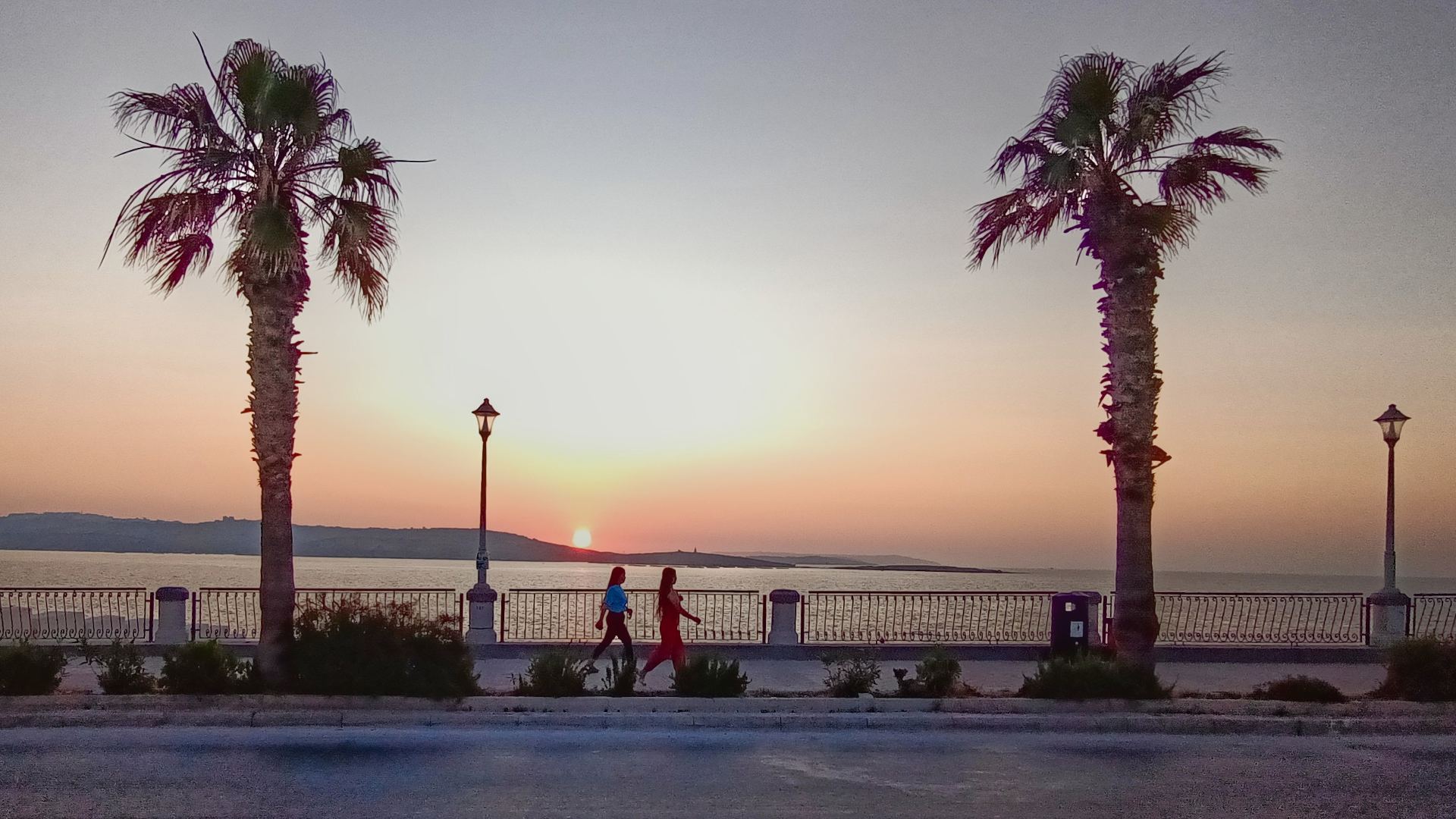 Sunset view on Malta: boulevard, sun is setting, two palm trees and two people walking by